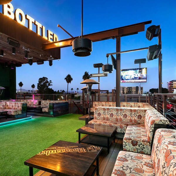 Rooftop bar with cushioned seating, wooden tables, and a large screen.
