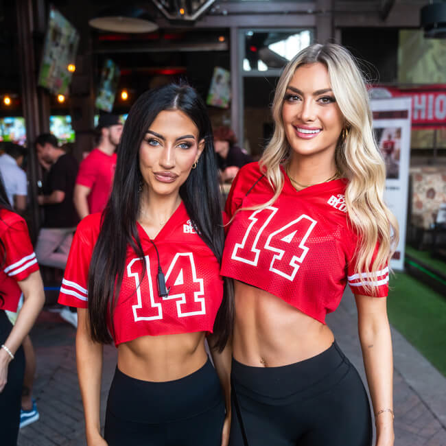 Two grils wearing matching red sports jerseys, standing together at a lively event.