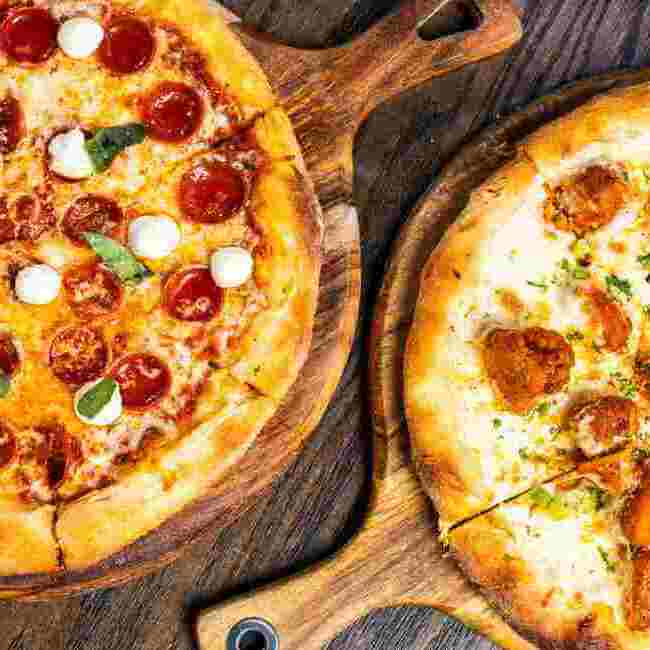 Overhead view of two freshly baked pizzas on wooden paddles, one topped with pepperoni, basil, and mozzarella balls, and the other with crispy chicken bites on a white sauce base.