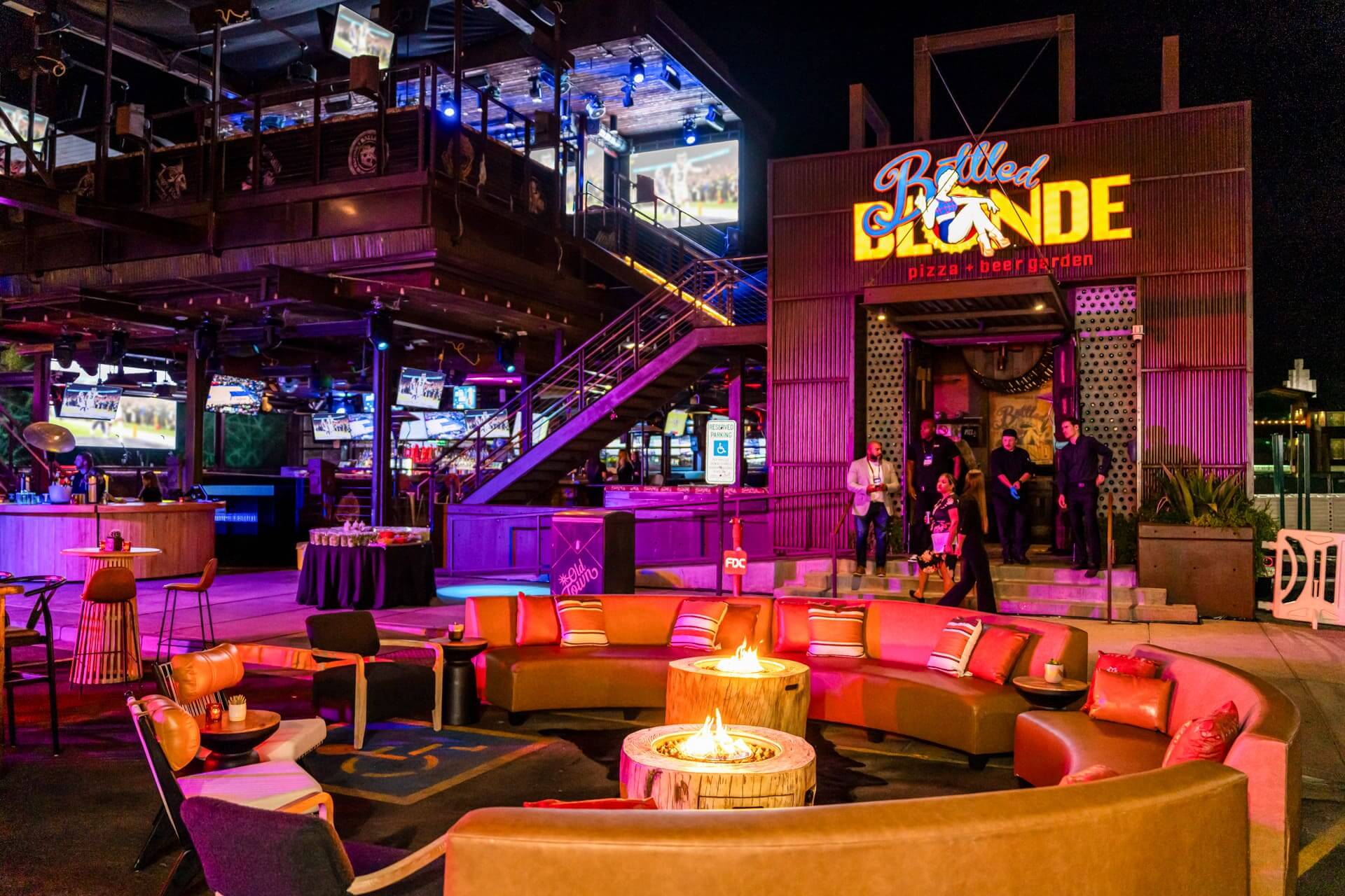 Outdoor lounge at Bottled Blonde featuring a circular firepit, modern seating, and neon lighting at night.