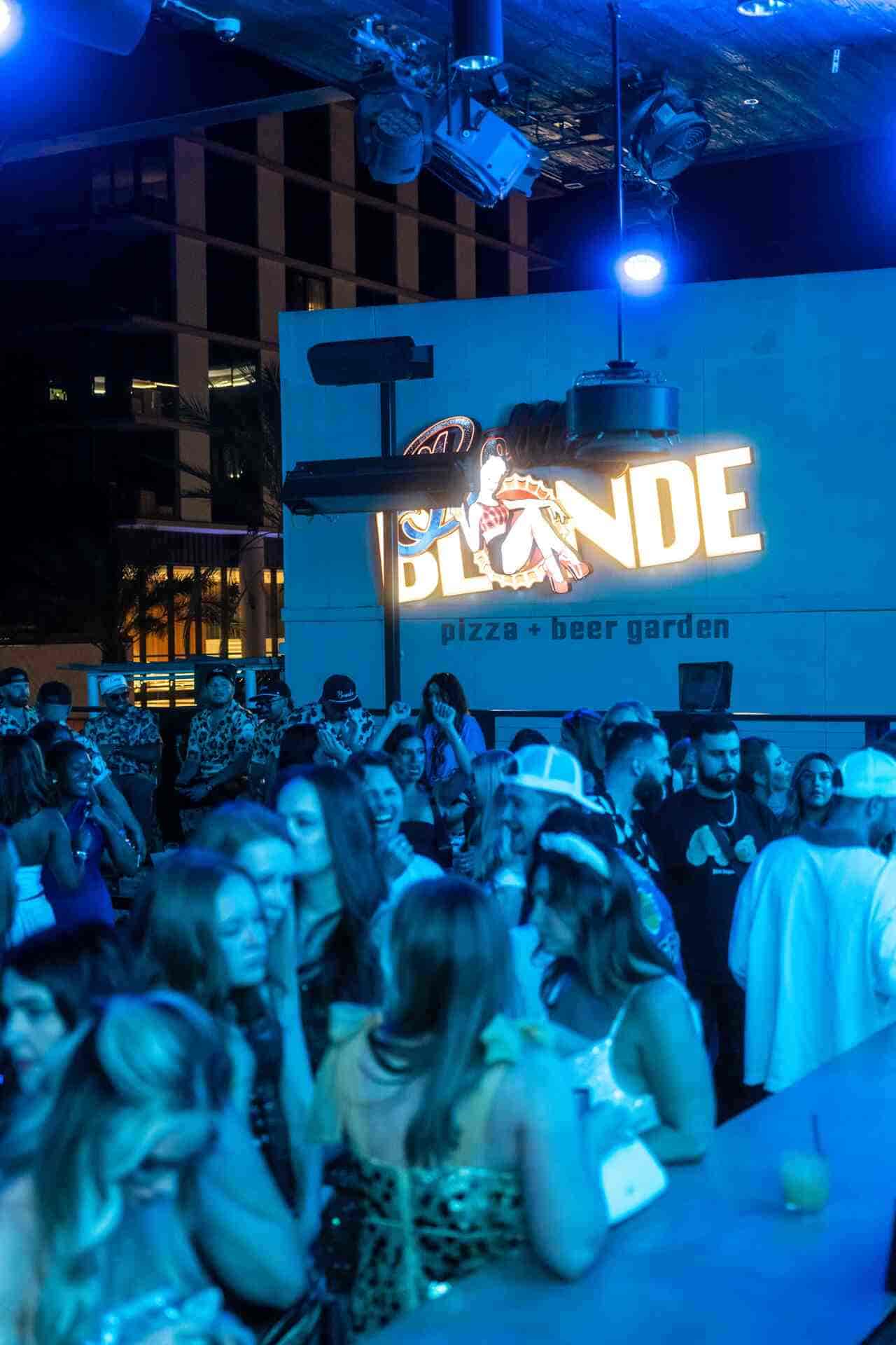 Packed rooftop party at Bottled Blonde beer garden under blue lights, with guests in costume.