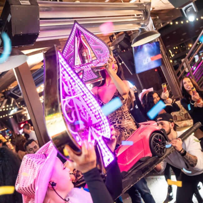 Nightlife crowd celebrating as performers hold illuminated ace-shaped sign on a pink scooter