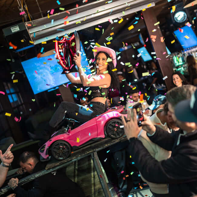 Bottle service performer riding a small pink car display while confetti falls in a crowded nightclub.