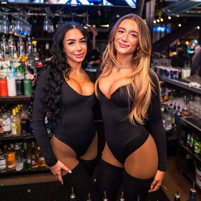 Two bartenders in black outfits smiling behind a bar with bottles and glassware