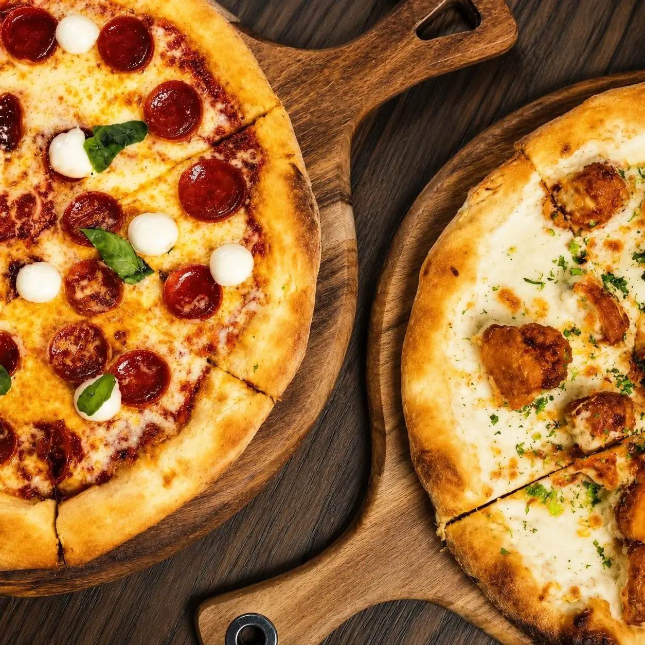 Overhead view of two artisan pizzas on wooden serving boards, featuring pepperoni, mozzarella, basil, and chicken toppings on a rustic wood table.