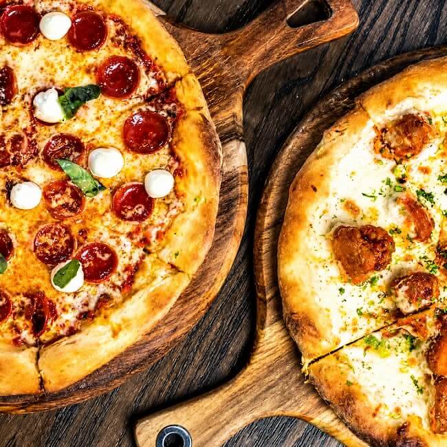 Overhead view of two artisan pizzas served on wooden boards, featuring a pepperoni pizza with mozzarella and basil and a chicken-topped white pizza on a rustic wood table.