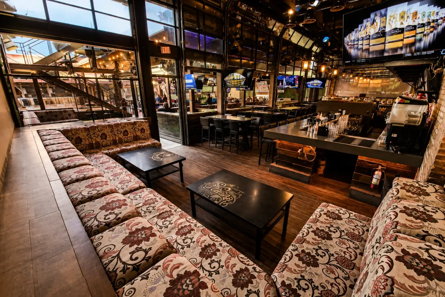 Spacious lounge seating area inside a modern bar featuring patterned couches, coffee tables, large televisions, and floor-to-ceiling windows overlooking the venue.