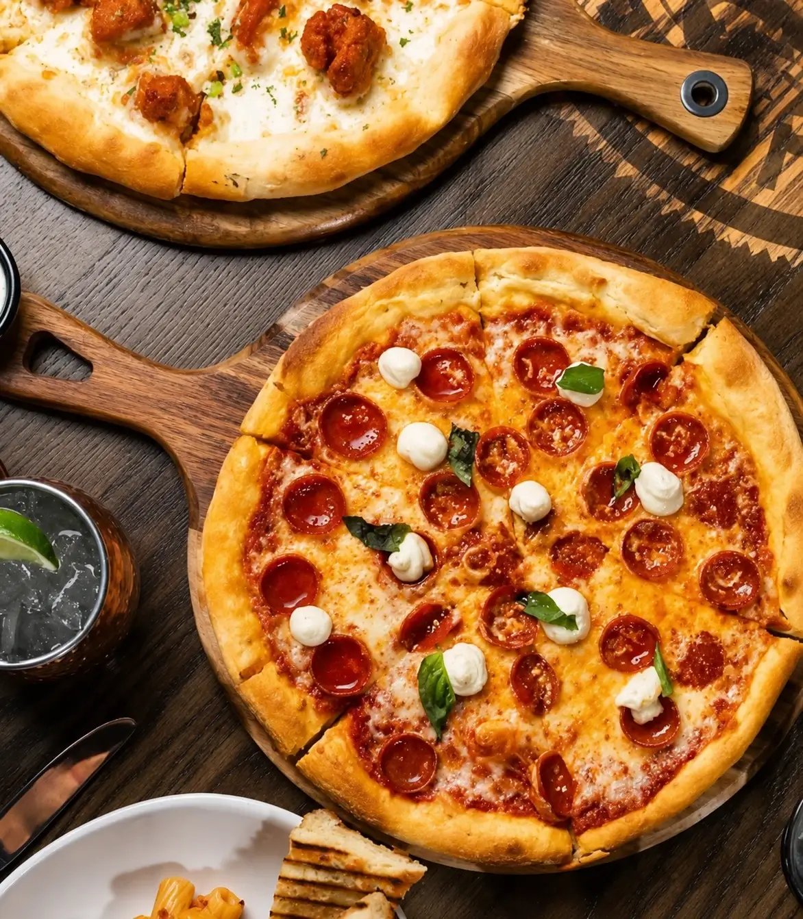 Overhead view of a pepperoni pizza topped with mozzarella and basil on a wooden serving board, surrounded by other dishes and drinks on a rustic table.